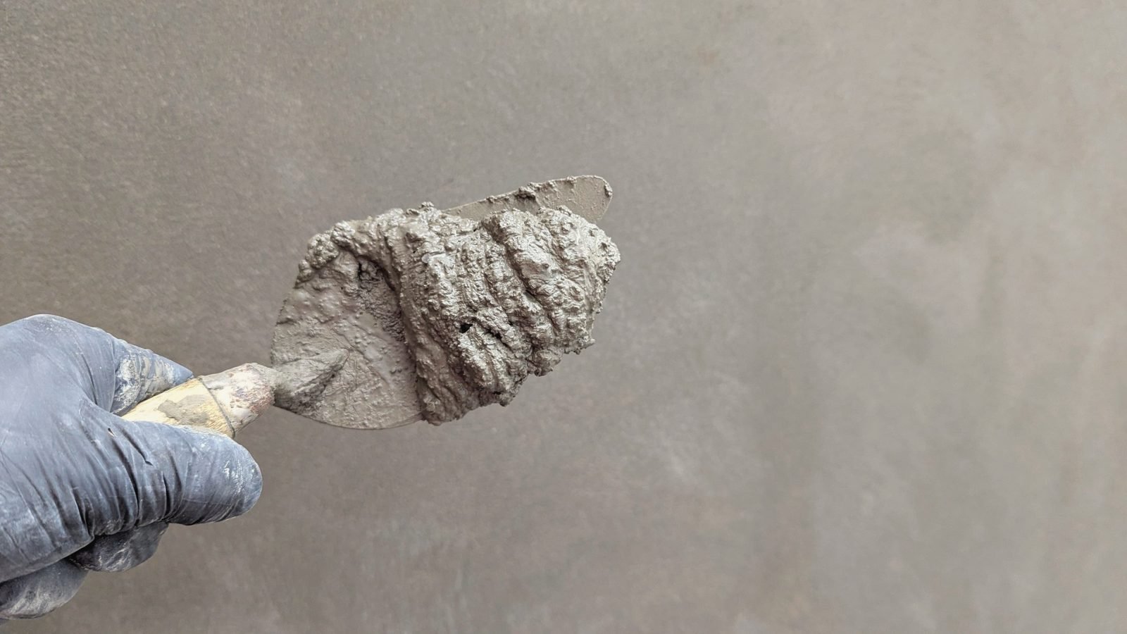A gloved hand holding a trowel of fresh cement against a smooth finished surface.